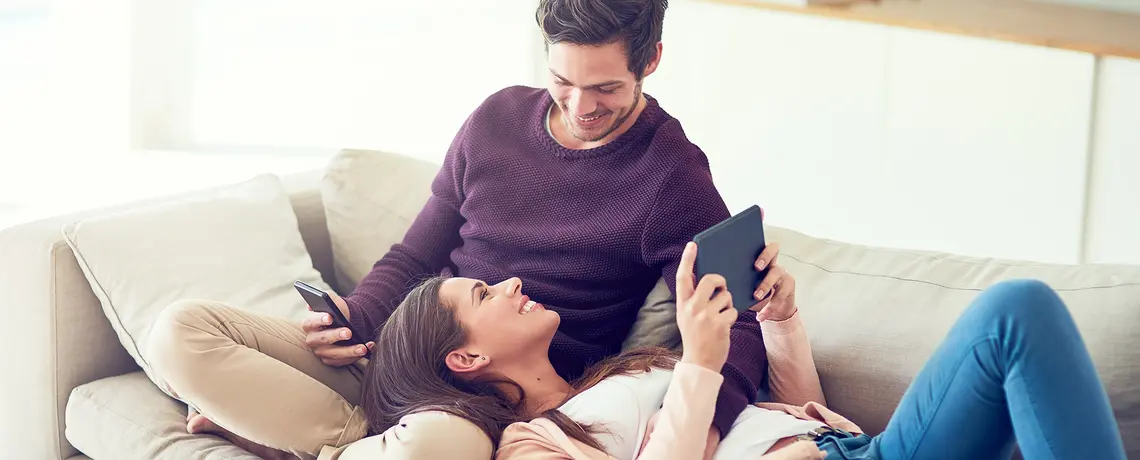 Eine Frau und ein Mann liegen auf dem Sofa. Er hat ein Smartphone in der Hand, sie ein Tablet.